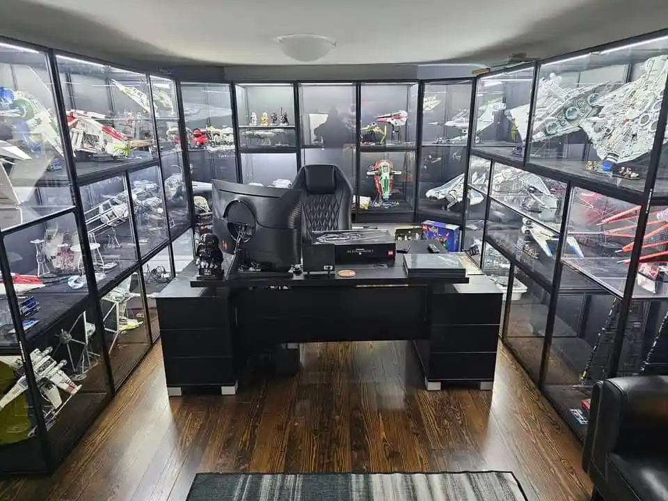 A room with a black desk and gaming chair centered, surrounded by tall glass display cases filled with Star Wars model ships…