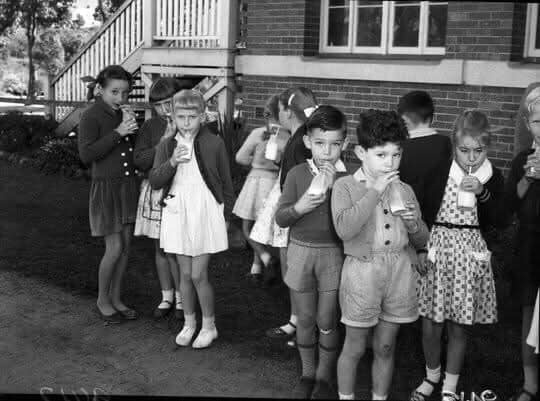 A black-and-white photo of several children in vintage school attire standing outdoors, each holding and eating a small…