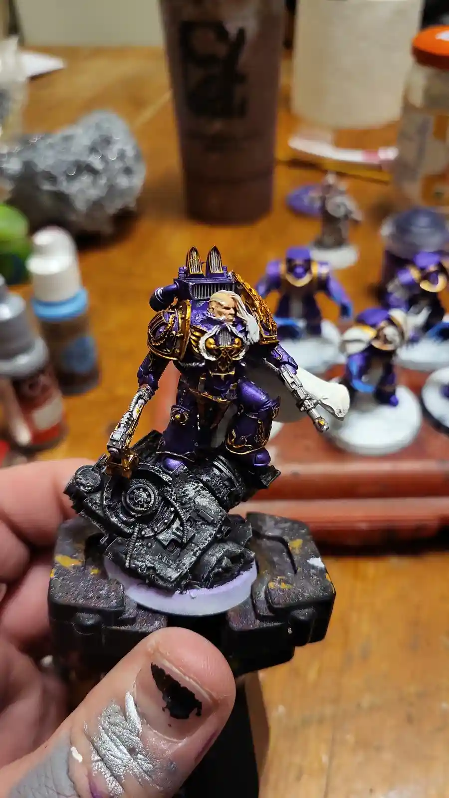 A close-up photo shows a hand holding a highly detailed, purple-and-gold painted miniature of a sci-fi warrior with a…