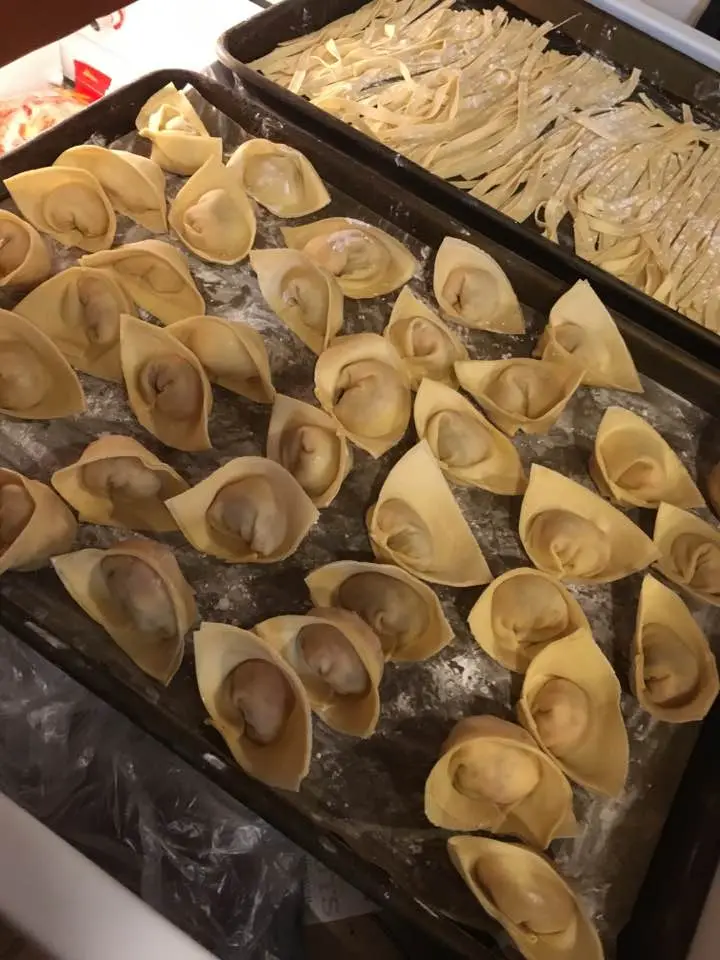 A close-up photo shows two metal trays filled with freshly made, uncooked dumplings and dumpling wrappers. The dumplings are…
