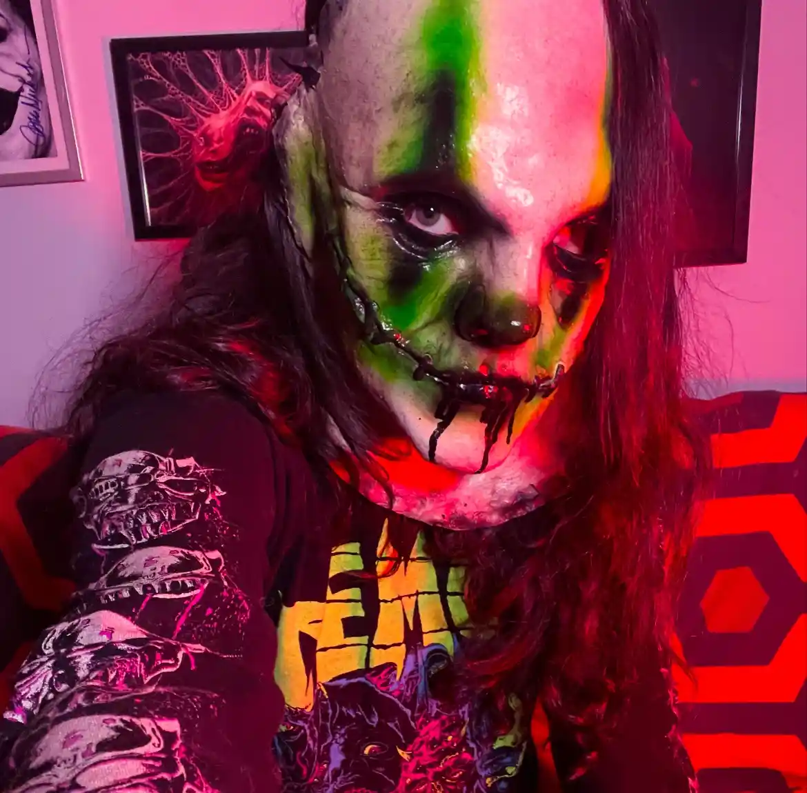 A person with long dark hair sits in a room lit with pink and red light, wearing a black t-shirt with graphic designs. Their…