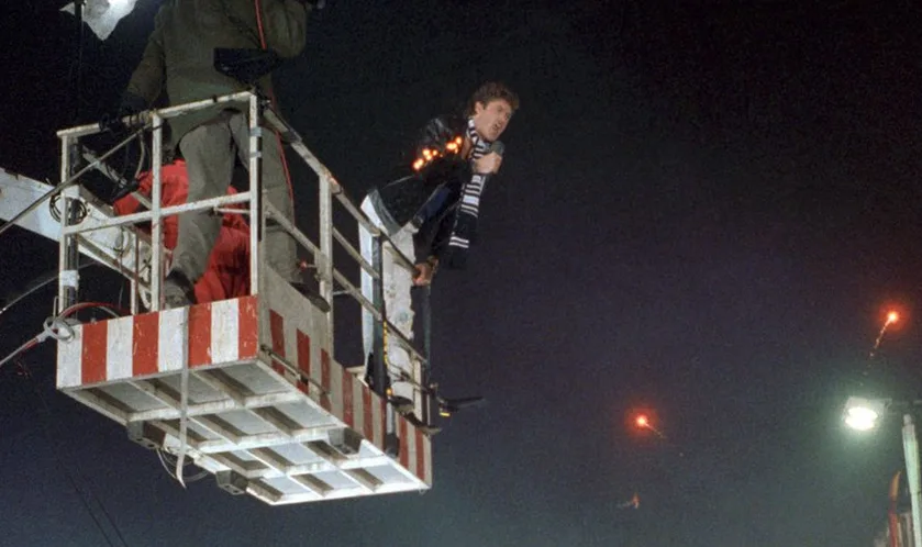 A man in a striped shirt and dark jacket stands on the edge of a red-and-white striped aerial lift platform at night…