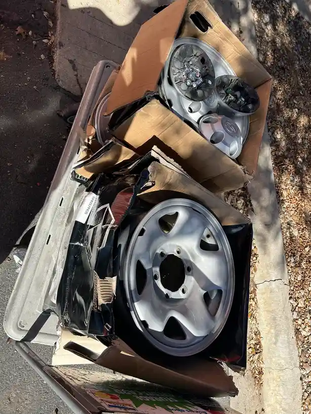 An overhead shot shows a metal trash bin overflowing with discarded car wheels and cardboard boxes on a sidewalk next to…