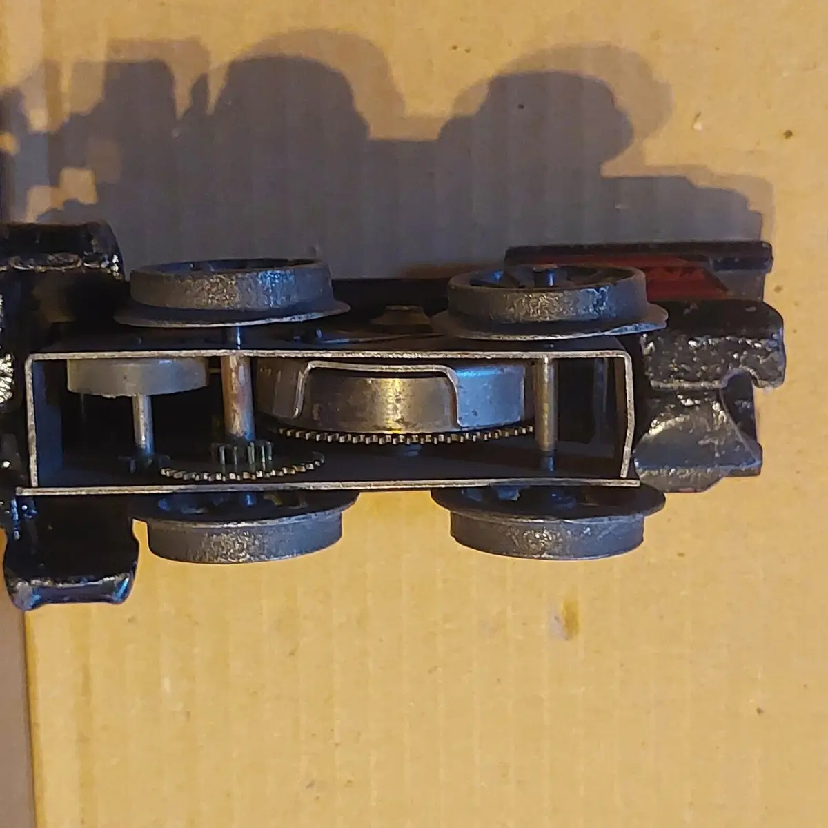 A close-up of a small, black metal train car with its undercarriage exposed, revealing wheels, axles, and gears. The shadow…