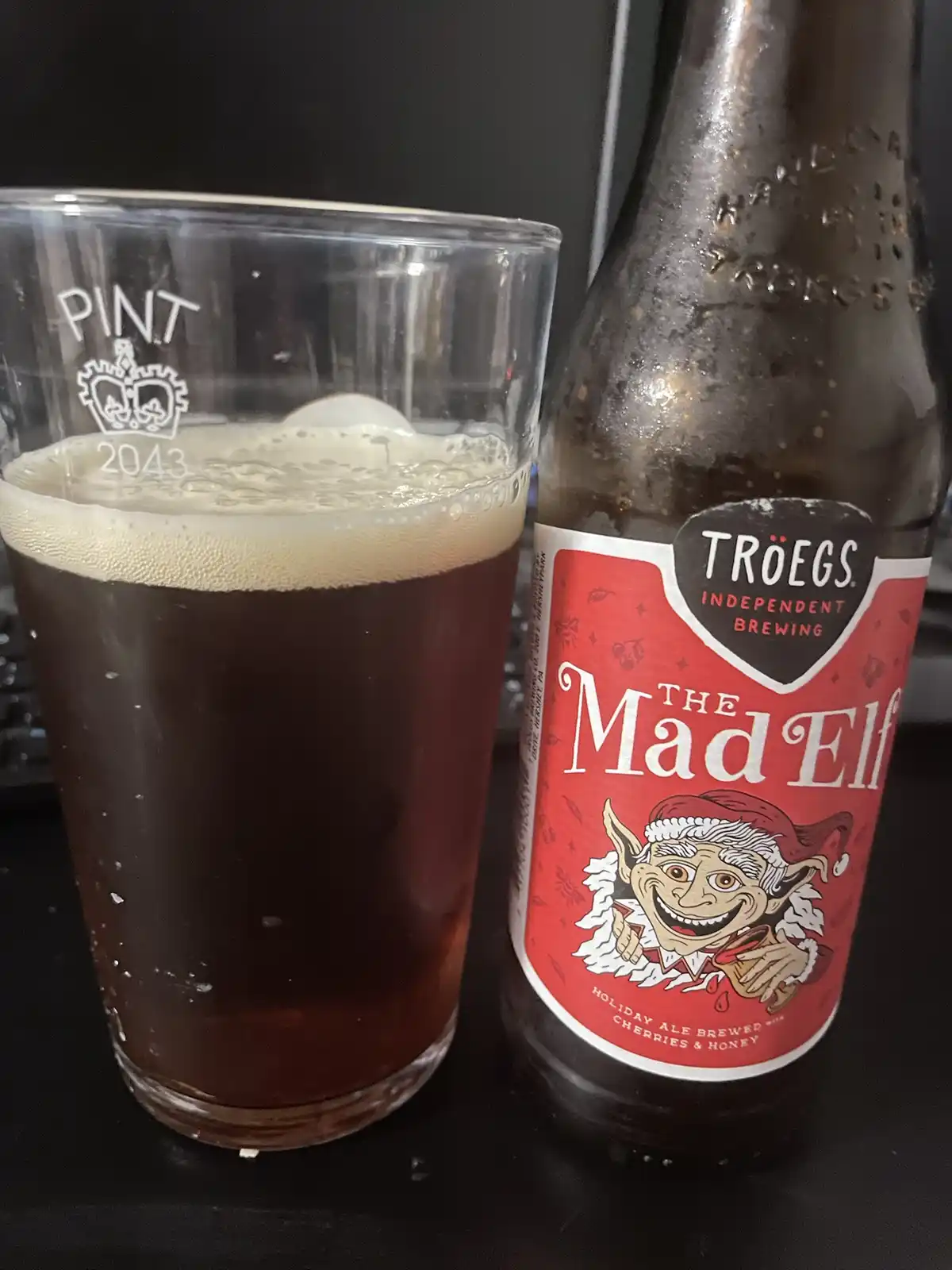 A pint glass filled with dark beer and a frothy head sits next to a chilled bottle of Troegs 'The Mad Elf' holiday ale…