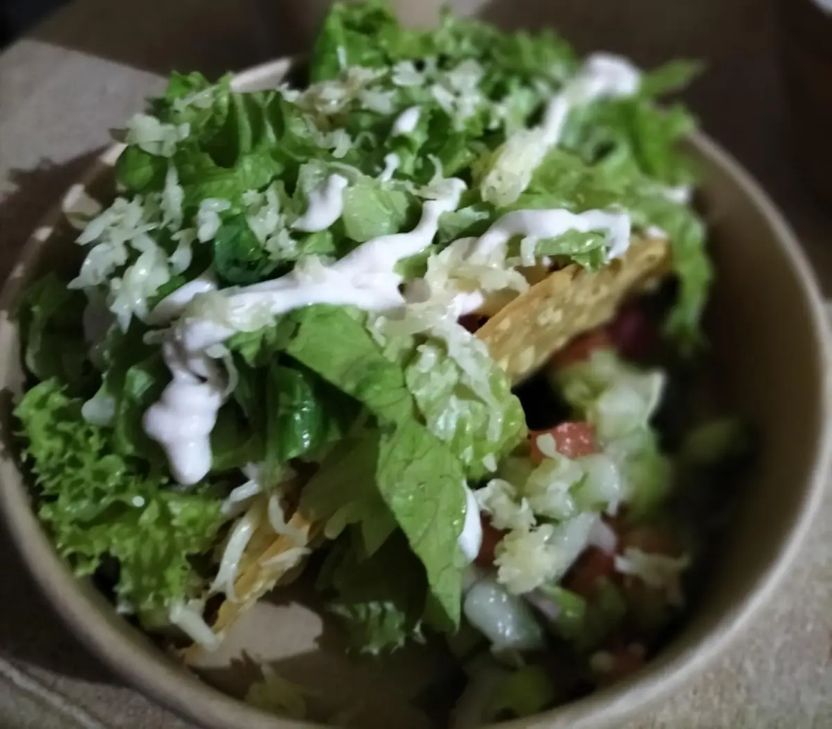 A close-up photo of a taco salad in a brown paper bowl, topped with shredded lettuce, shredded cheese, and a white sauce…