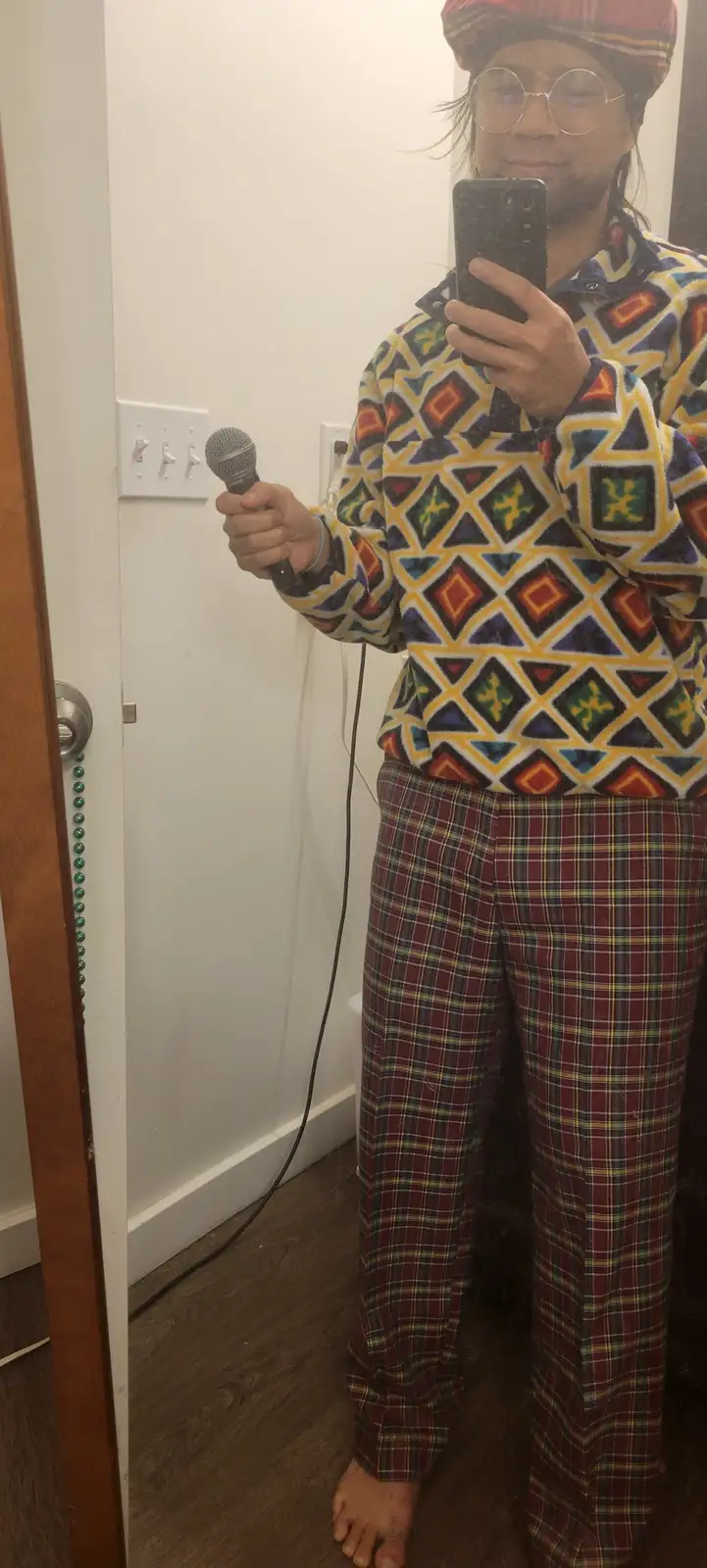 A person wearing a colorful geometric-patterned fleece shirt and plaid pajama pants stands barefoot in front of a mirror…