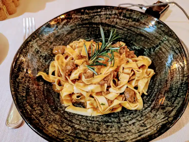 A close-up photo of a dark, textured ceramic plate holding wide pasta noodles mixed with meat sauce and garnished with a…