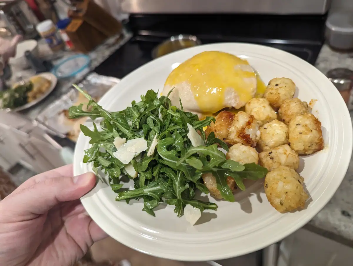 A hand holds a white plate with a meal consisting of a poached egg with hollandaise sauce, roasted tater tots, and a side of…