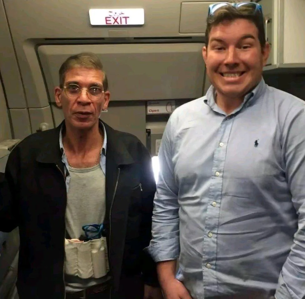 Two men stand side by side inside an airplane cabin near an exit sign. The man on the left, wearing glasses and a black…