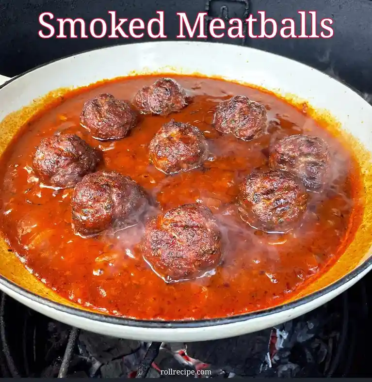 A white enamel pan sits over glowing charcoal briquettes, containing nine browned meatballs simmering in a bubbling, red…