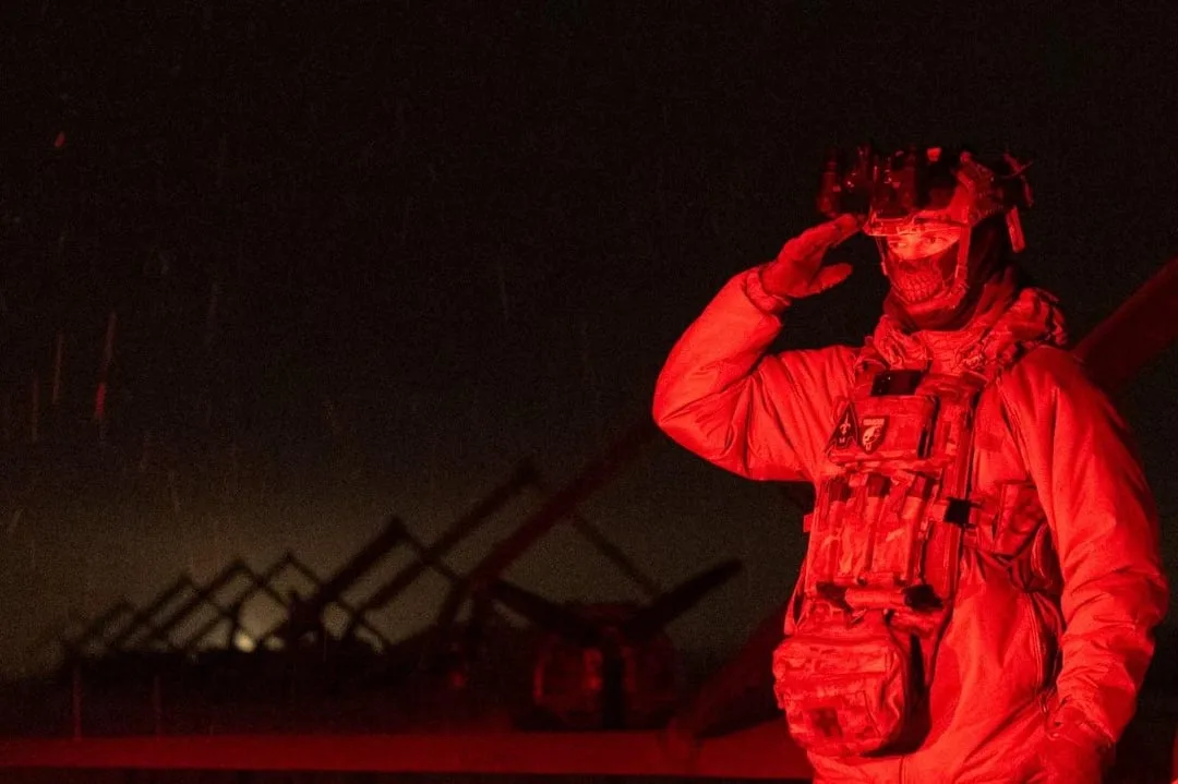 A soldier in full tactical gear, including a helmet with night vision goggles and a face mask, salutes under red lighting at…