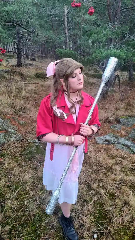 A person in a cosplay outfit with a pink bow, red jacket, and white dress stands in a forest holding a sword entirely…