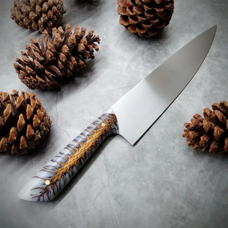 A chef's knife with a handle designed to look like a pinecone, surrounded by actual pinecones on a gray surface. The…