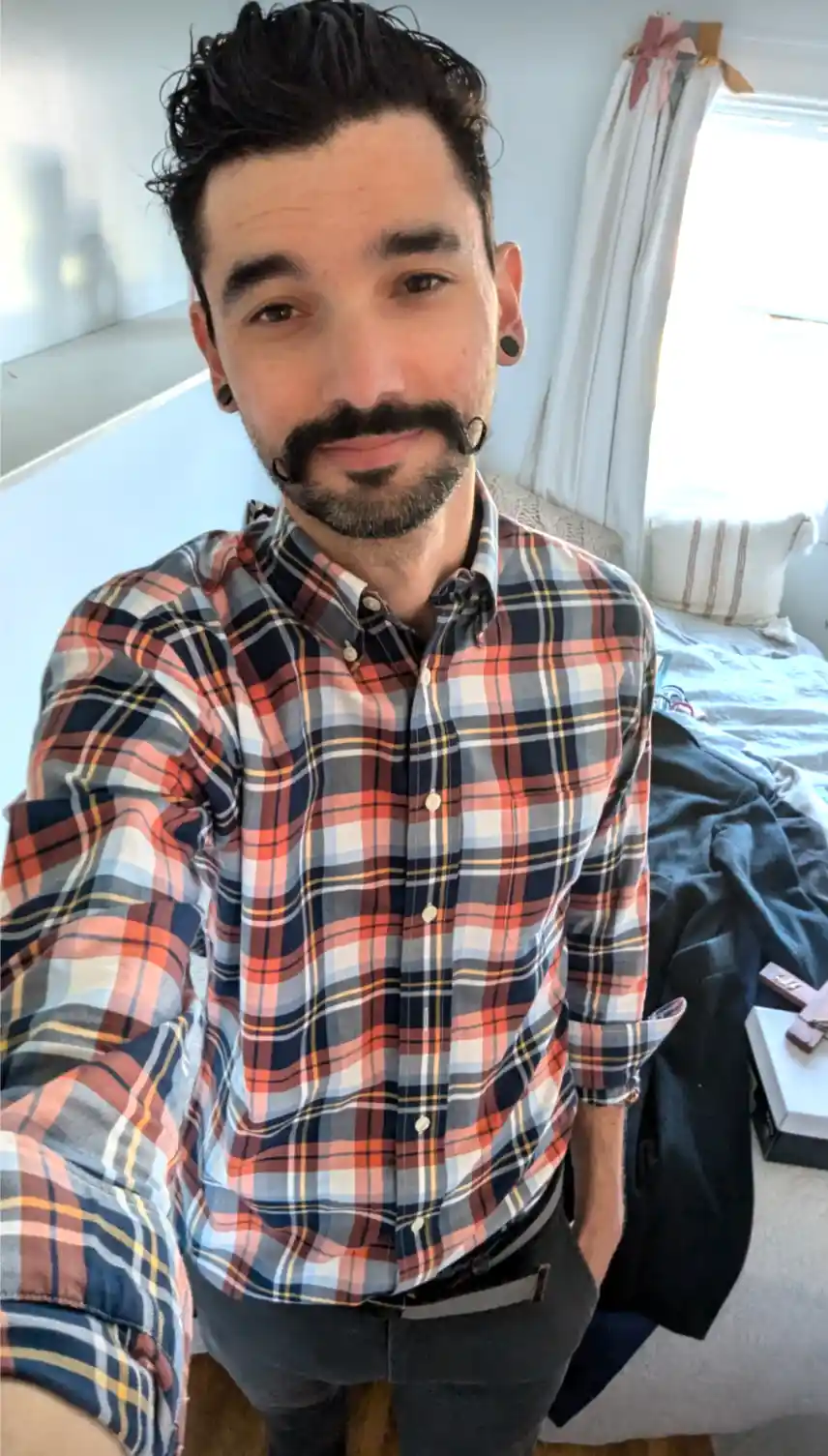 A man with a goatee and black ear gauges takes a selfie in a bedroom wearing a plaid shirt. The text overlaid on the image…