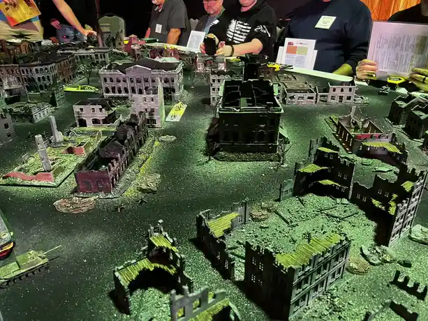 A tabletop wargame setup shows a cityscape of miniature, heavily damaged buildings and rubble under green lighting, with…