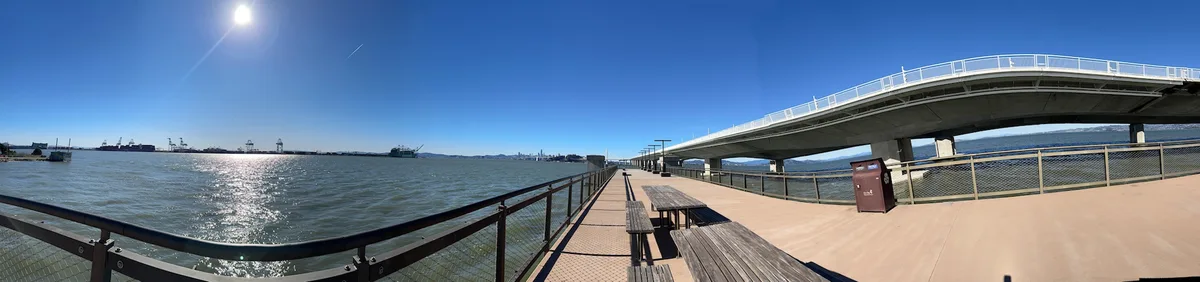 A wide-angle photo of a sunny waterfront promenade with picnic tables, railings, and a large bridge overhead. The sky is…