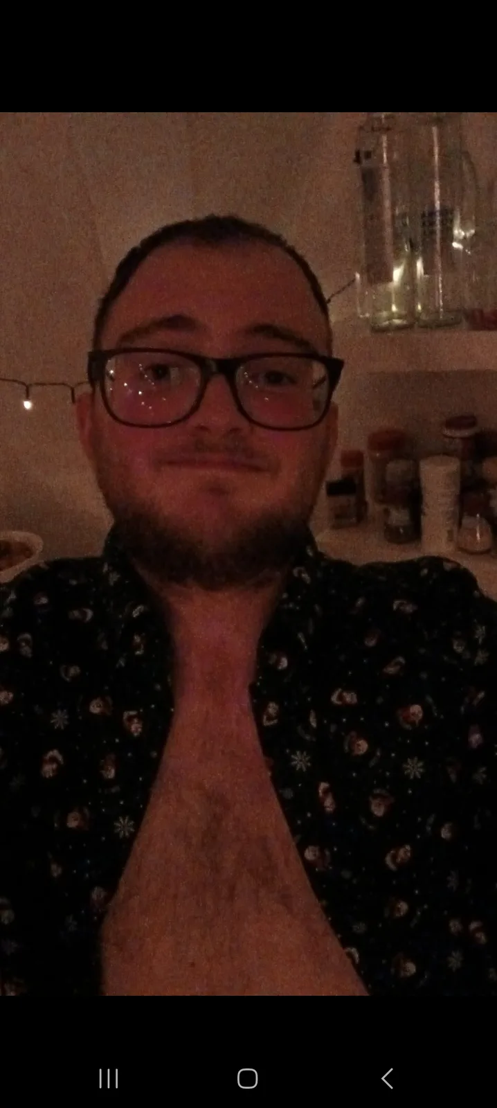 A man with glasses and a beard, wearing a dark shirt with a holiday pattern, looks at the camera with a slight smirk while…