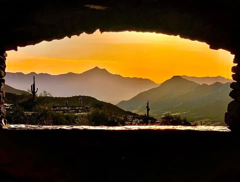 A scenic desert sunset viewed through a rough stone archway, with silhouetted mountains, cacti, and a warm orange sky. The…