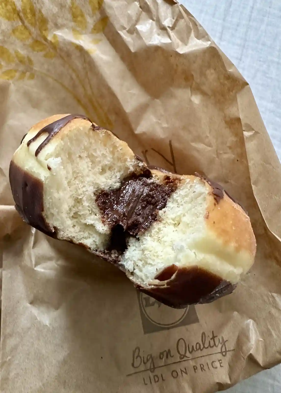 A close-up photo shows a partially eaten chocolate-drizzled pastry with a thick chocolate filling, resting on brown Lidl…