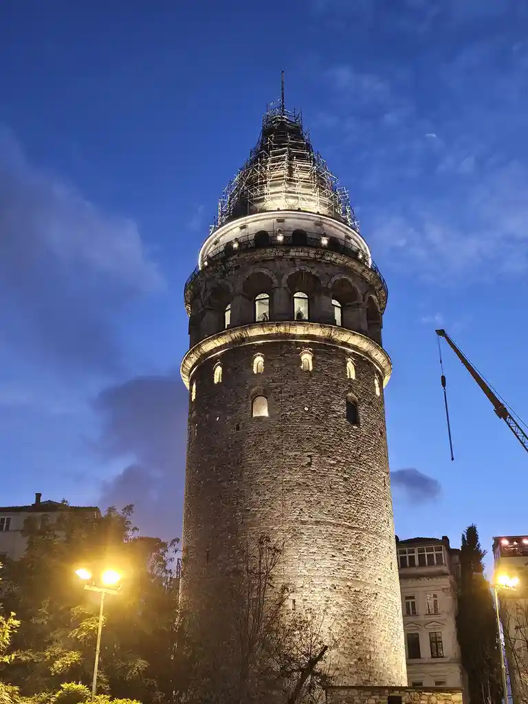 a low-angle nighttime photo shows the galata tower in istanbul, its upper section covered in scaffolding and illuminated…