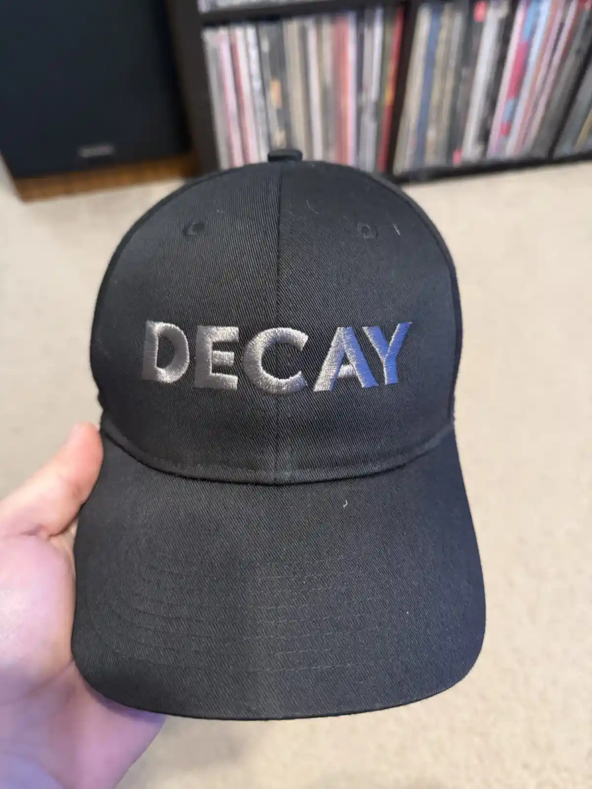 A hand holds a black baseball cap with the word 'DECAY' embroidered across the front in silver and blue thread. The…