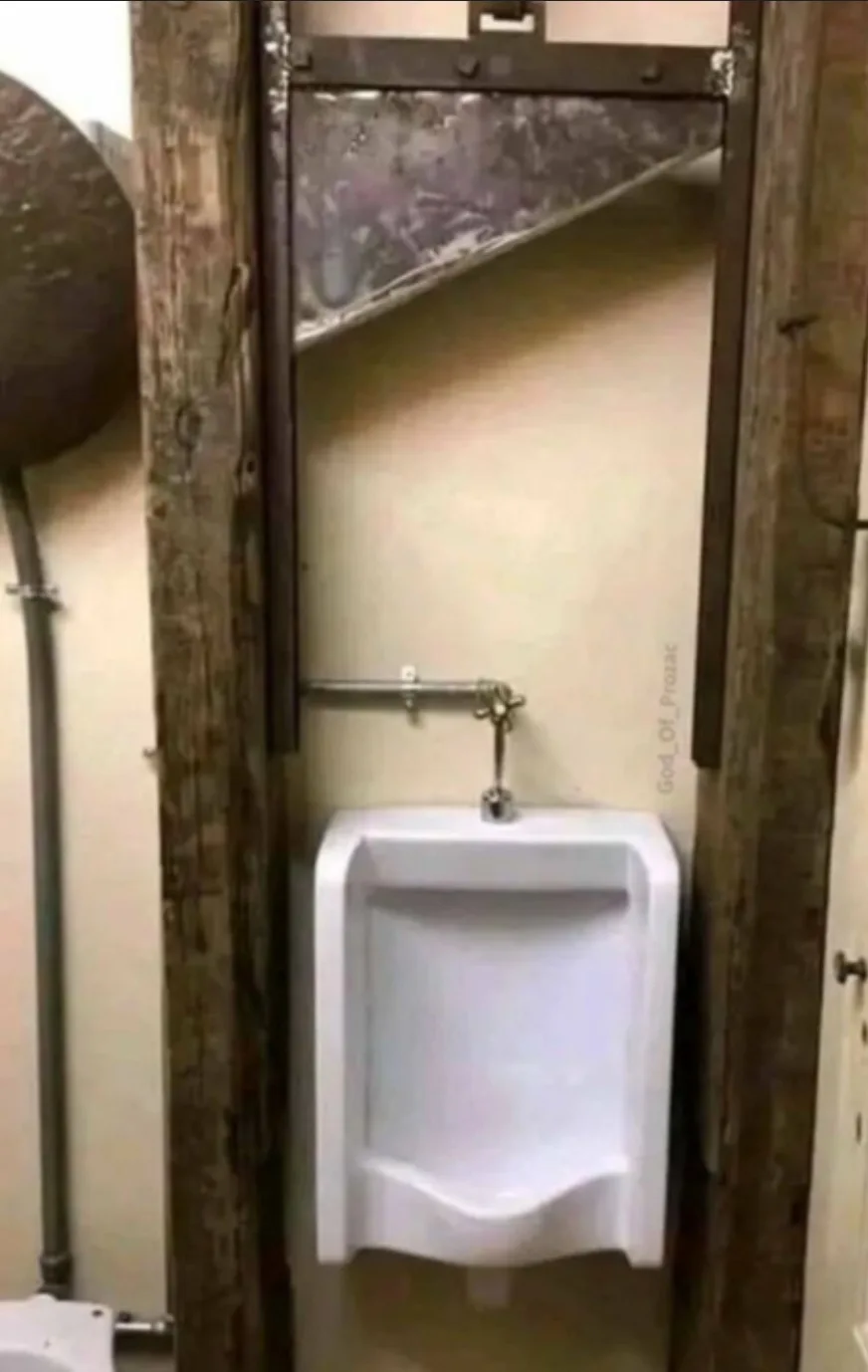 A white urinal is mounted on a beige wall inside a dilapidated bathroom stall framed by weathered, dark wooden beams. Above…