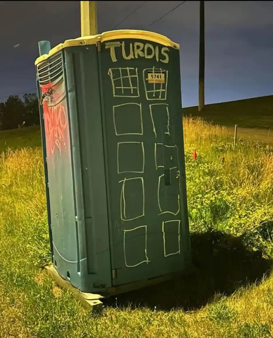 A green utility box, resembling a British telephone booth, sits in a grassy field with a dark sky. It has been crudely…