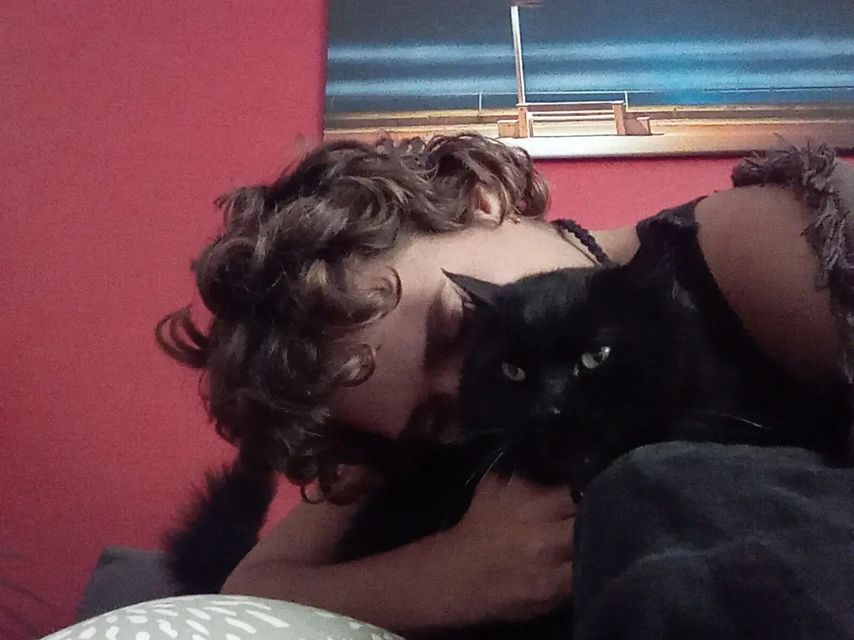 A person with curly brown hair lies on their side against a red wall, cuddling a black cat that looks directly at the camera…
