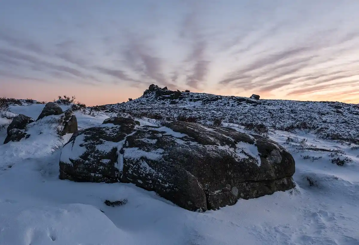 A serene, snow-covered rocky landscape at dawn or dusk, with large boulders in the foreground and a distant rocky ridge…