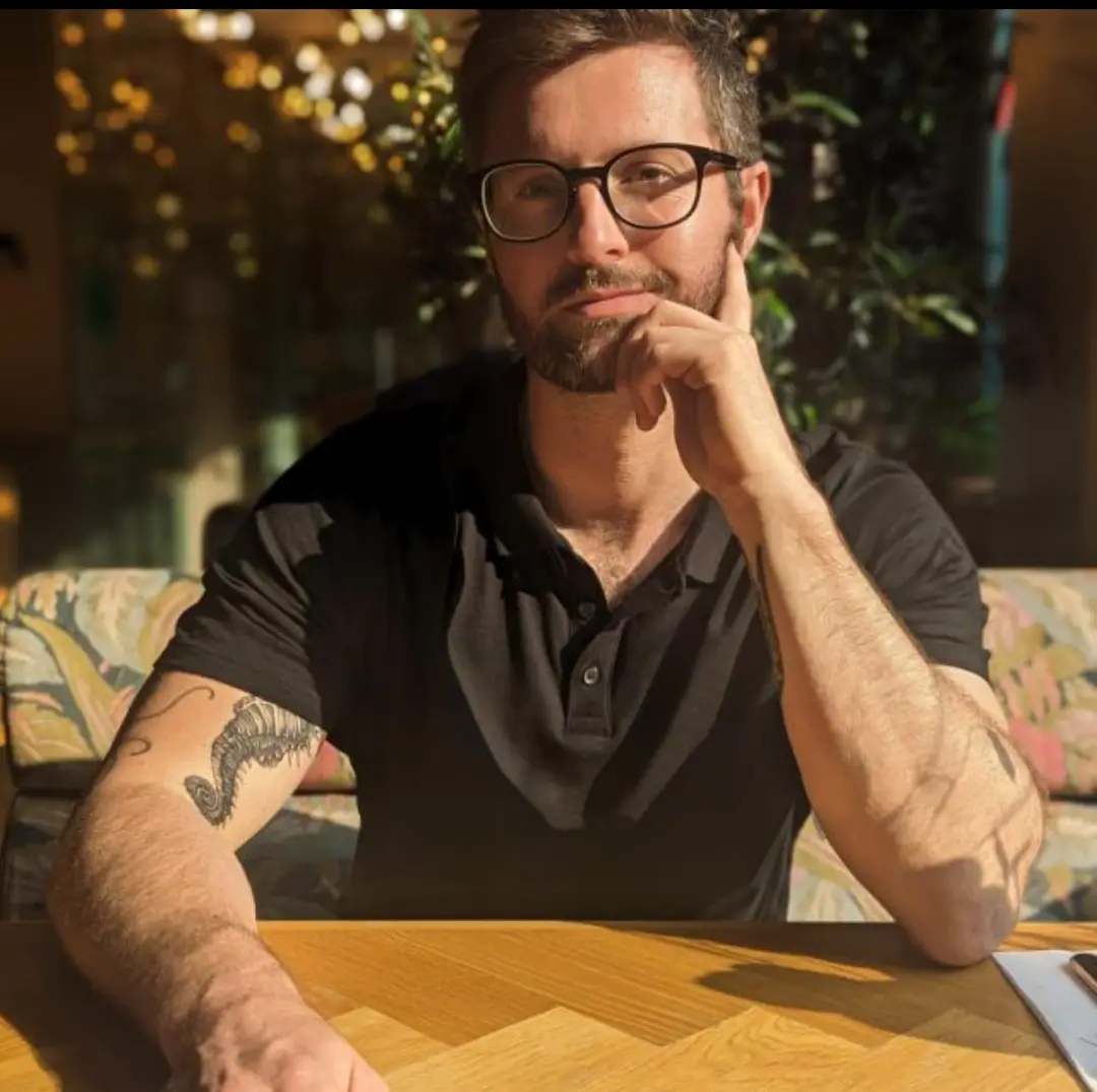 a man with glasses and a beard sits at a wooden table, resting his chin on his hand in a thoughtful pose. He wears a black…