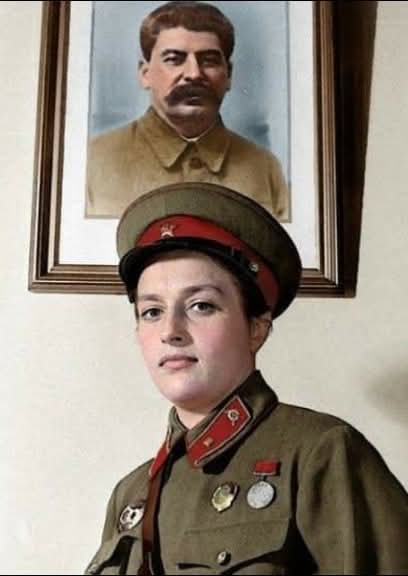 A young woman in a Soviet military uniform with medals stands in front of a framed portrait of Joseph Stalin. The overlaid…
