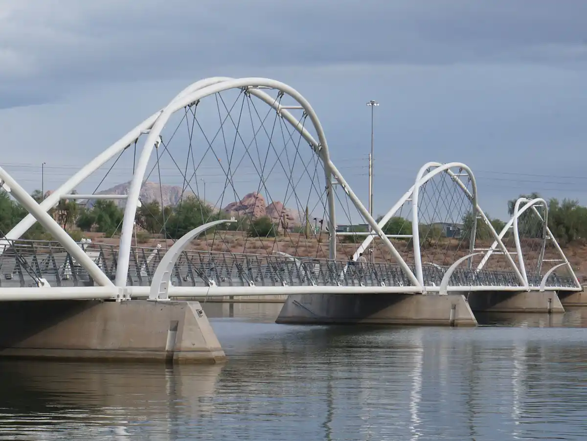 A photograph of a modern white arched pedestrian bridge spanning calm water, with desert hills and sparse vegetation in the…