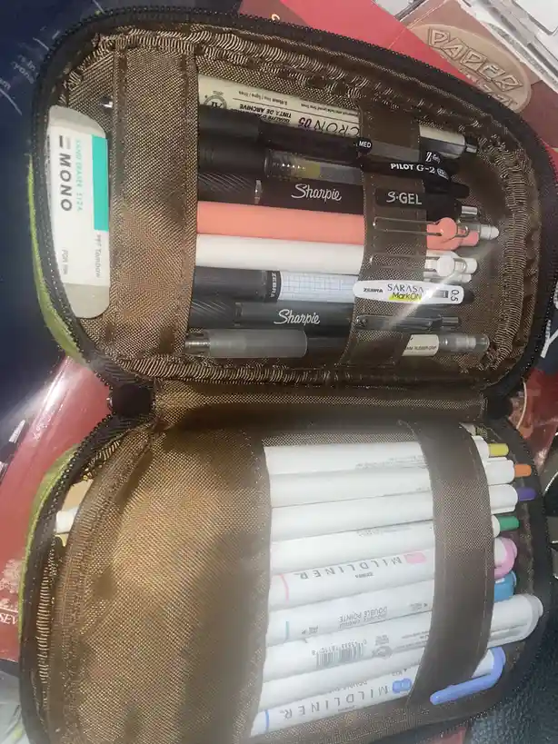 An open brown fabric pencil case filled with numerous pens, markers, and an eraser, neatly organized in compartments. The…