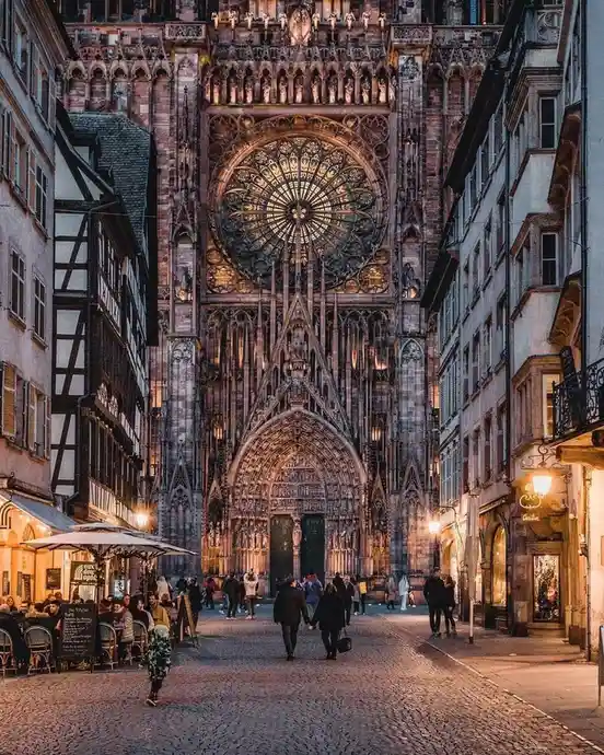 A wide-angle photo shows a cobblestone street flanked by buildings, leading to the grand, illuminated facade of a Gothic…