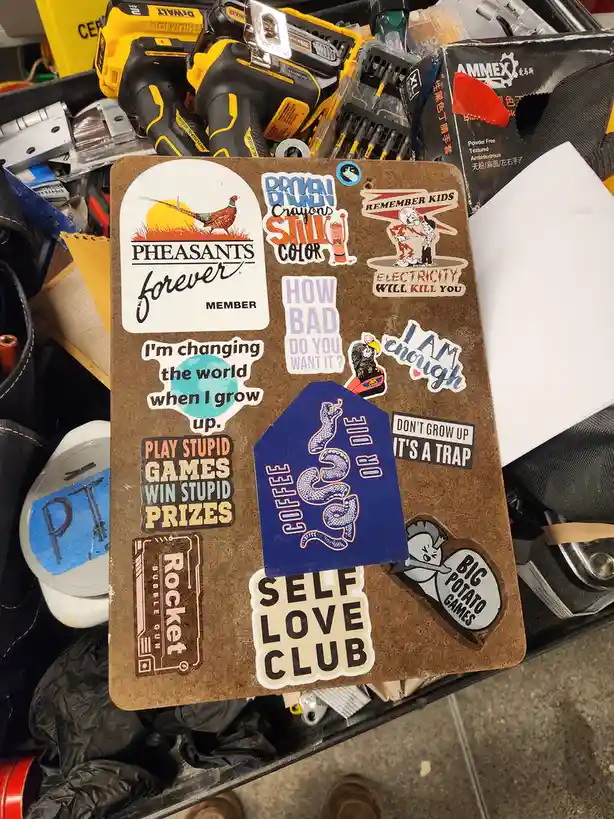A brown cork board covered in various stickers, including 'Pheasants Forever Member', 'Broken Crayons Still Color', 'I'm…