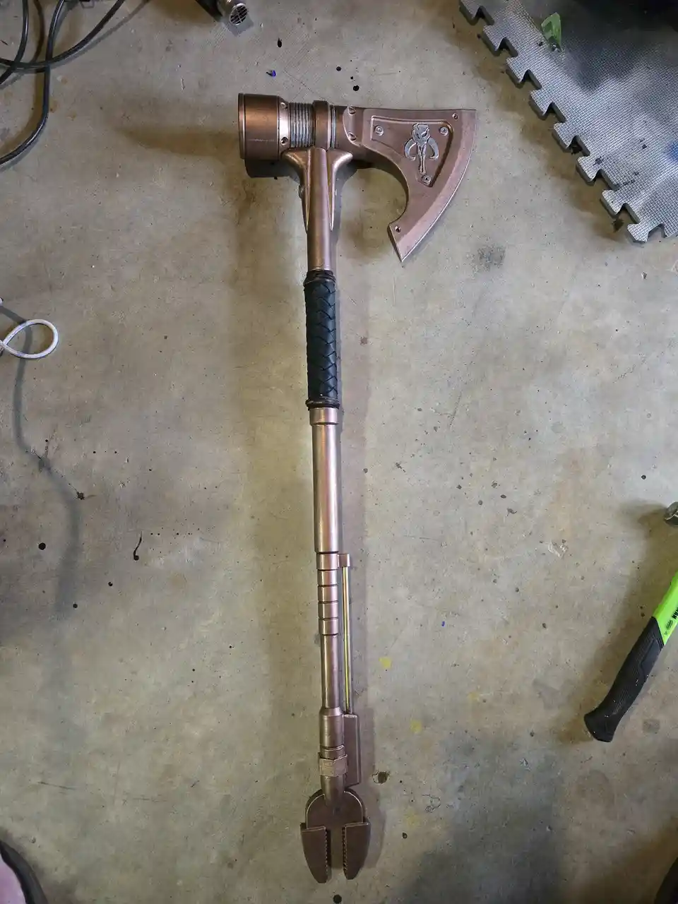 A custom-made axe with a copper-colored metal head featuring a Mandalorian symbol, a black braided grip, and a long shaft…