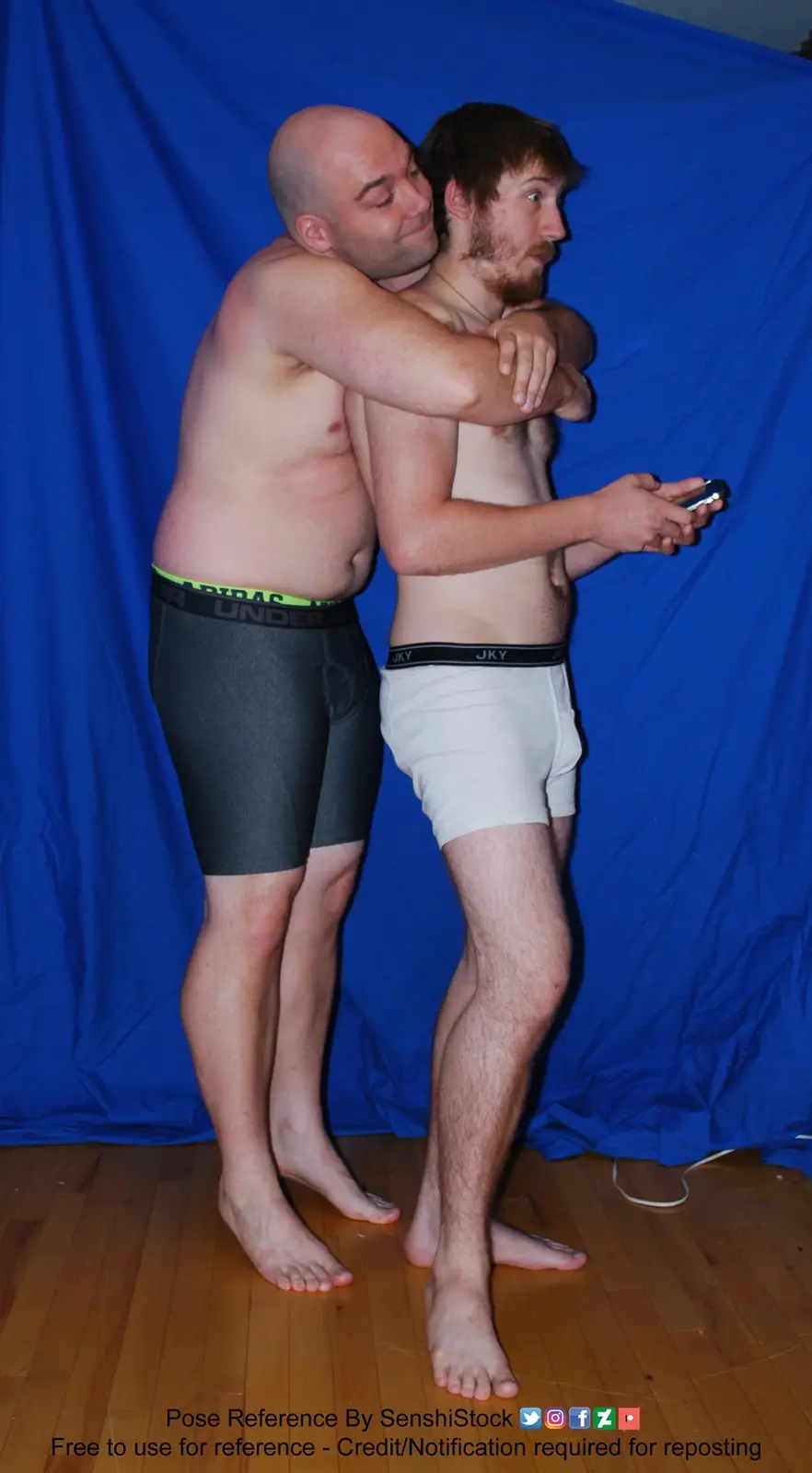 Two shirtless men stand barefoot on a wooden floor against a blue backdrop, one embracing the other from behind while the…