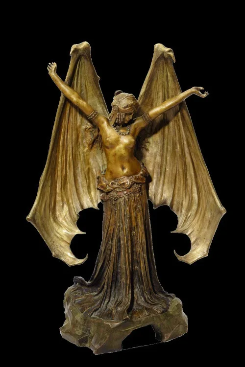 A bronze statue of a female figure with large, bat-like wings spread wide, arms raised, standing on a rocky base against a…