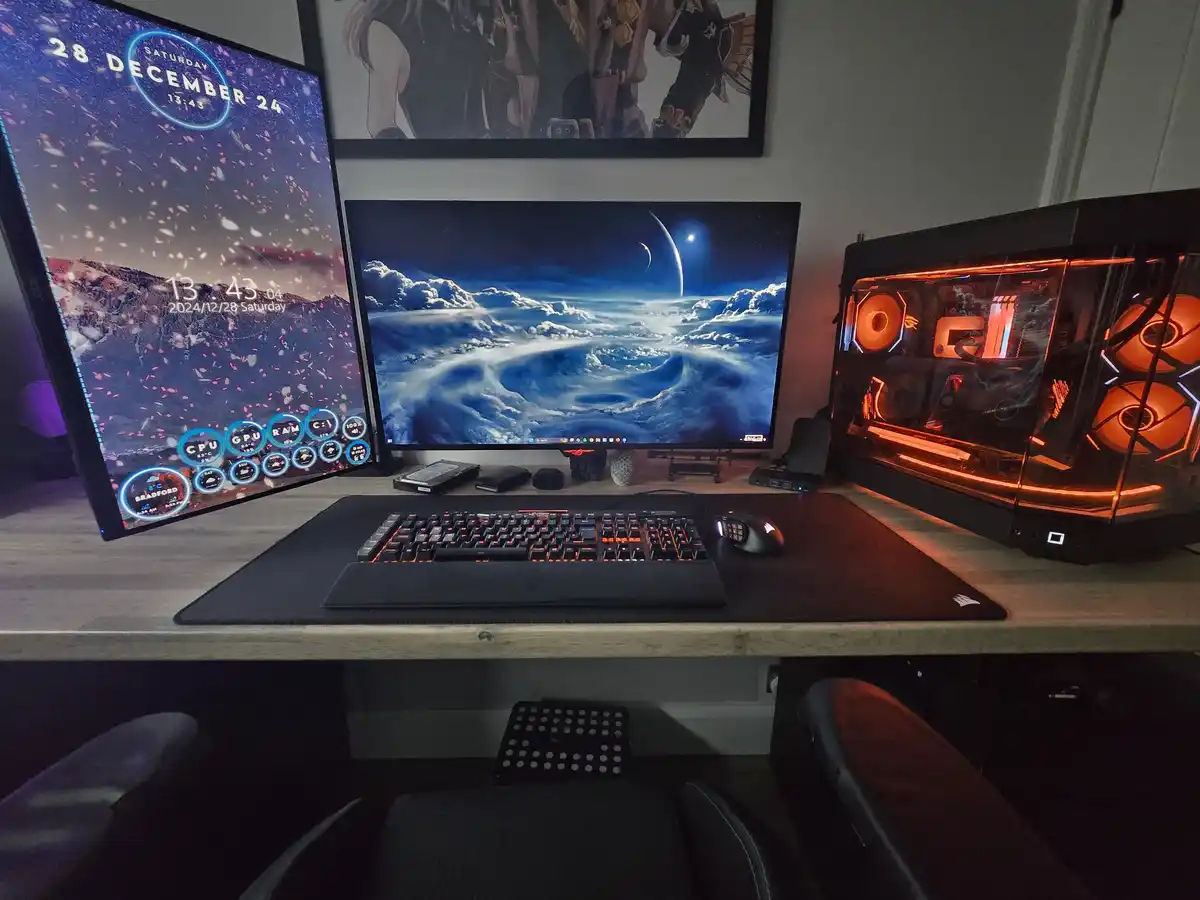 A computer desk setup with two monitors, a glowing orange PC case, and a keyboard mouse on a large mat. The left monitor…