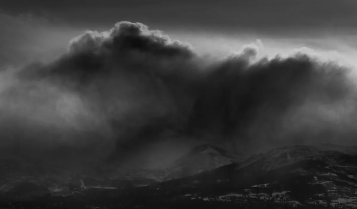 A black-and-white photograph shows dramatic, dark storm clouds looming over snow-dusted mountains and a valley. The overlaid…