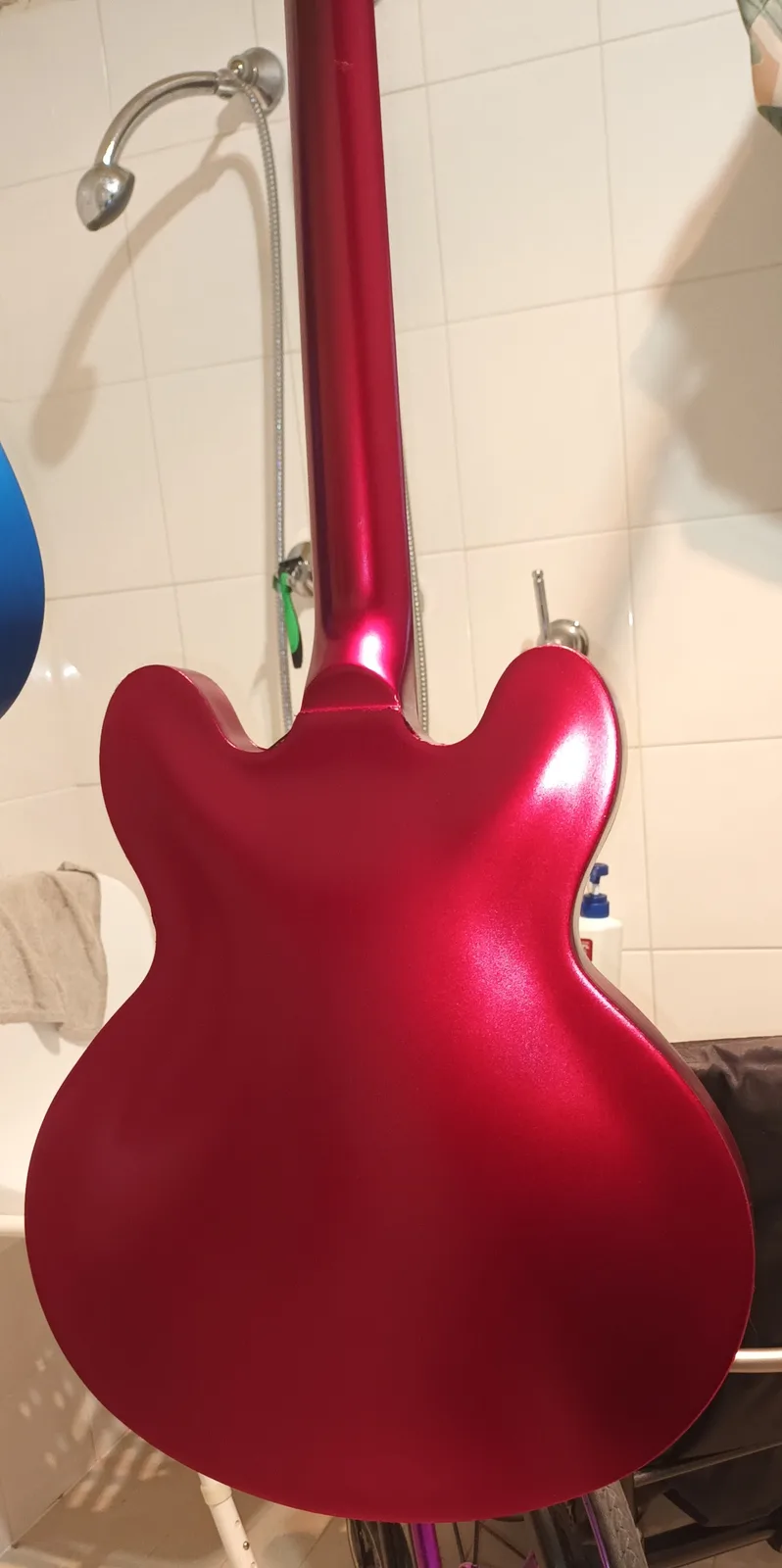 A shiny red electric guitar is standing upright in a bathroom, its back facing the camera, positioned directly in front of a…