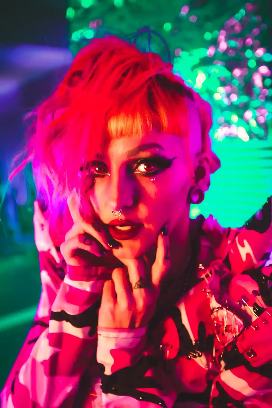 A person with short, brightly colored hair and heavy makeup poses with hands near their face against a neon-lit background…