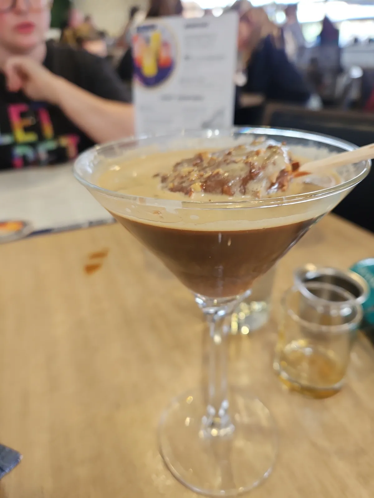A close-up photo shows a martini glass filled with a layered beverage, dark brown at the bottom and lighter beige on top…