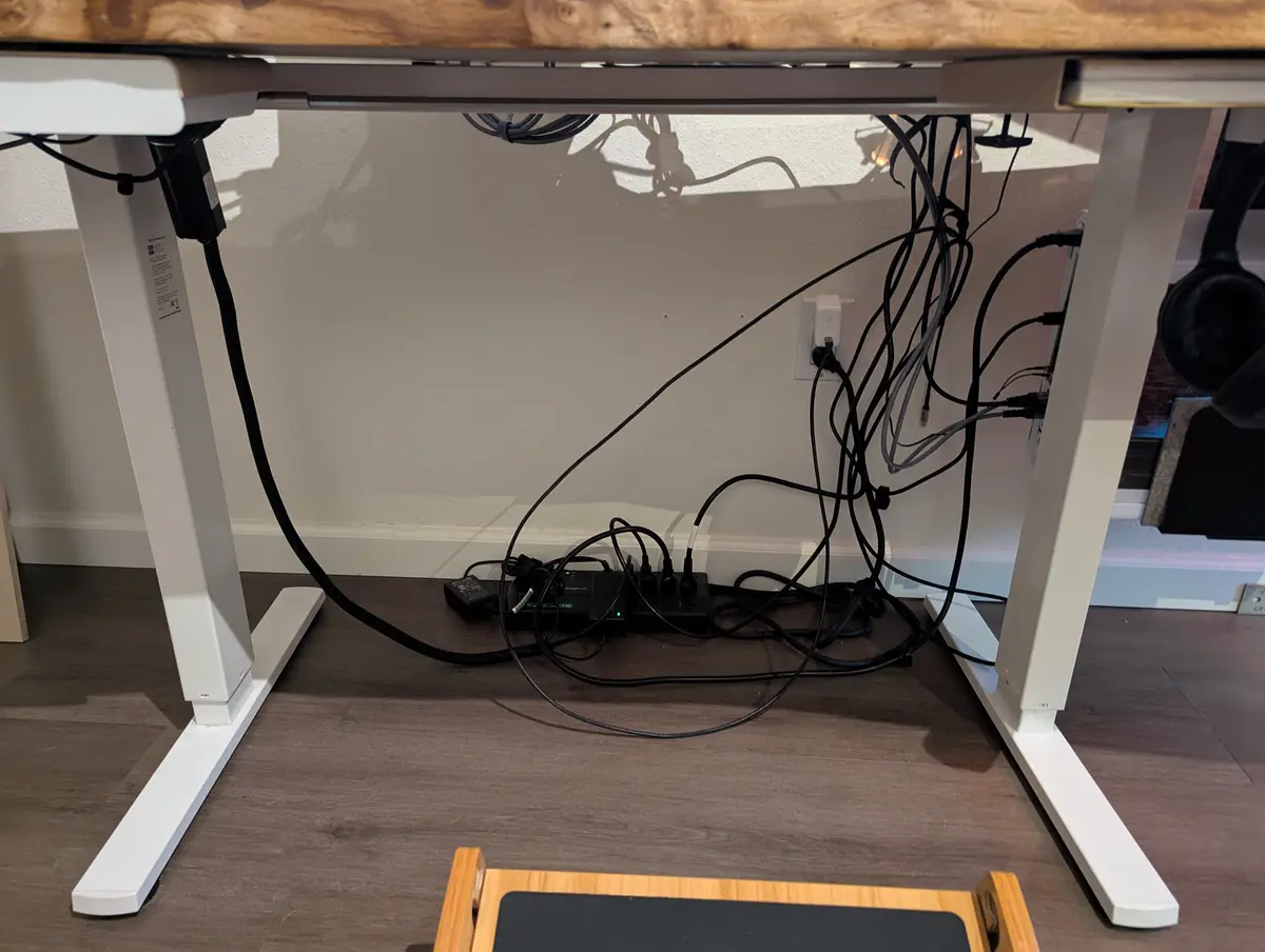 A photo shows the underside of a modern white standing desk with a wooden top, revealing a chaotic tangle of black power…