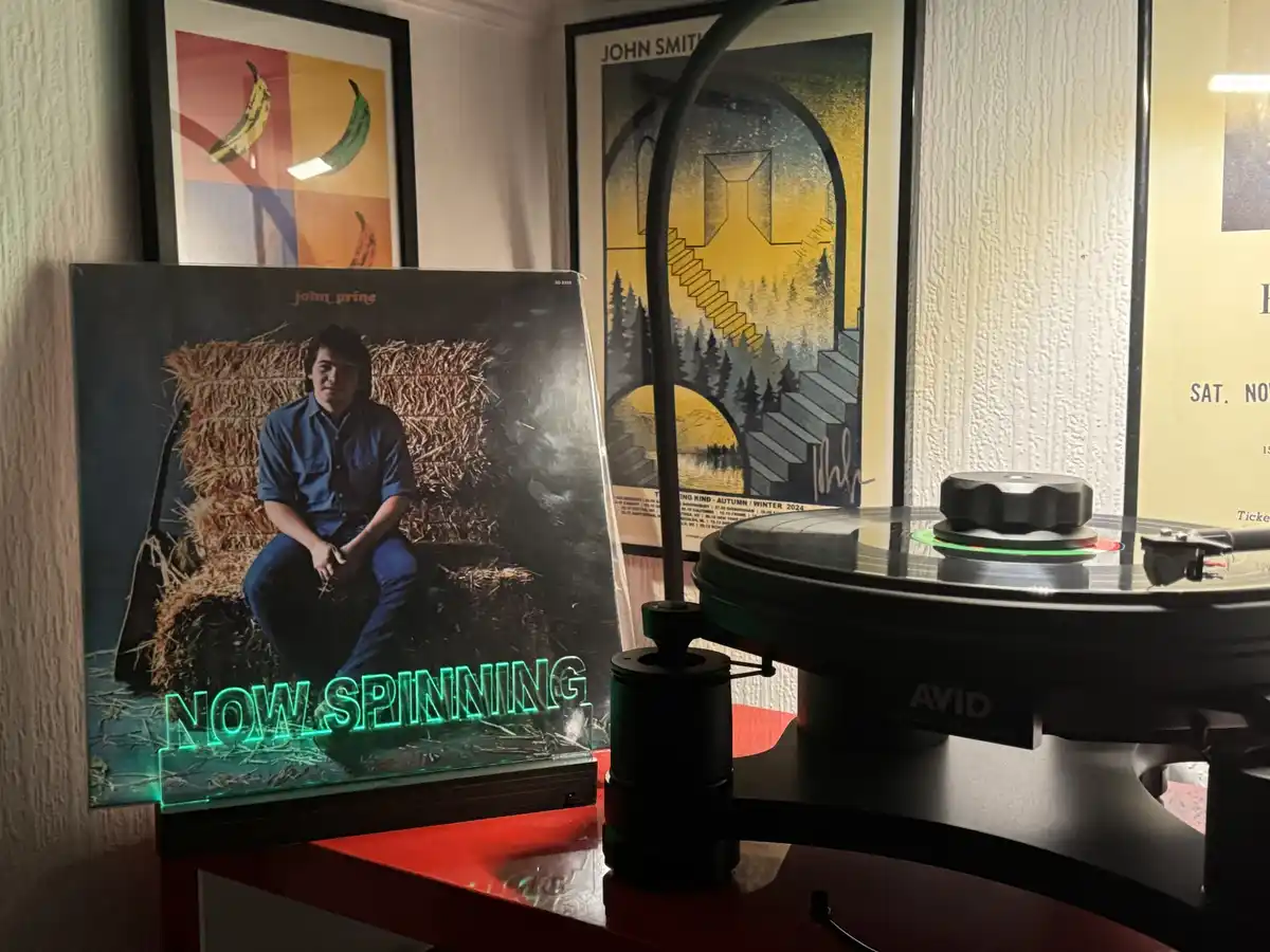 A vinyl record player with a glowing green 'NOW SPINNING' sign sits next to the album cover of John Prine, which shows him…