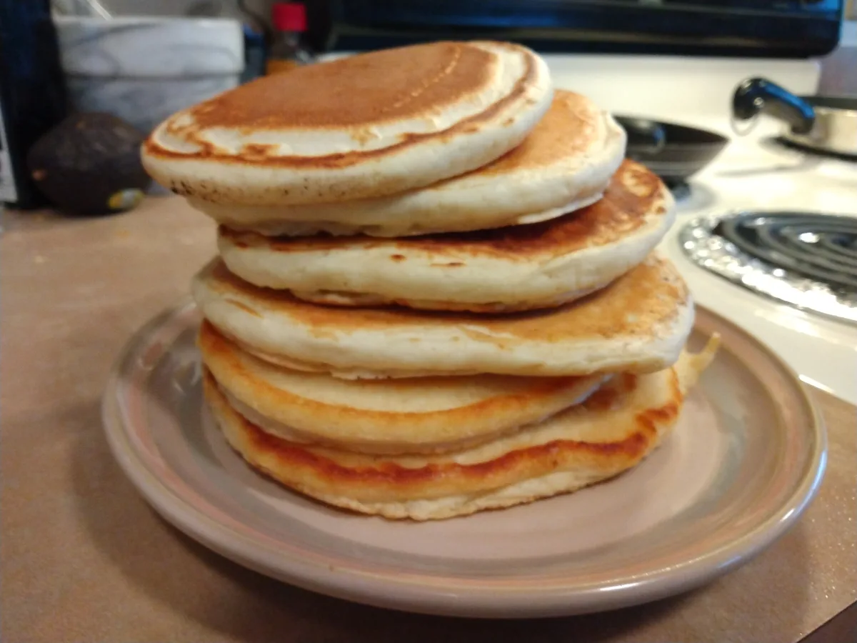 A tall stack of golden-brown pancakes sits on a light-colored plate on a kitchen counter, with a stove and avocado visible…