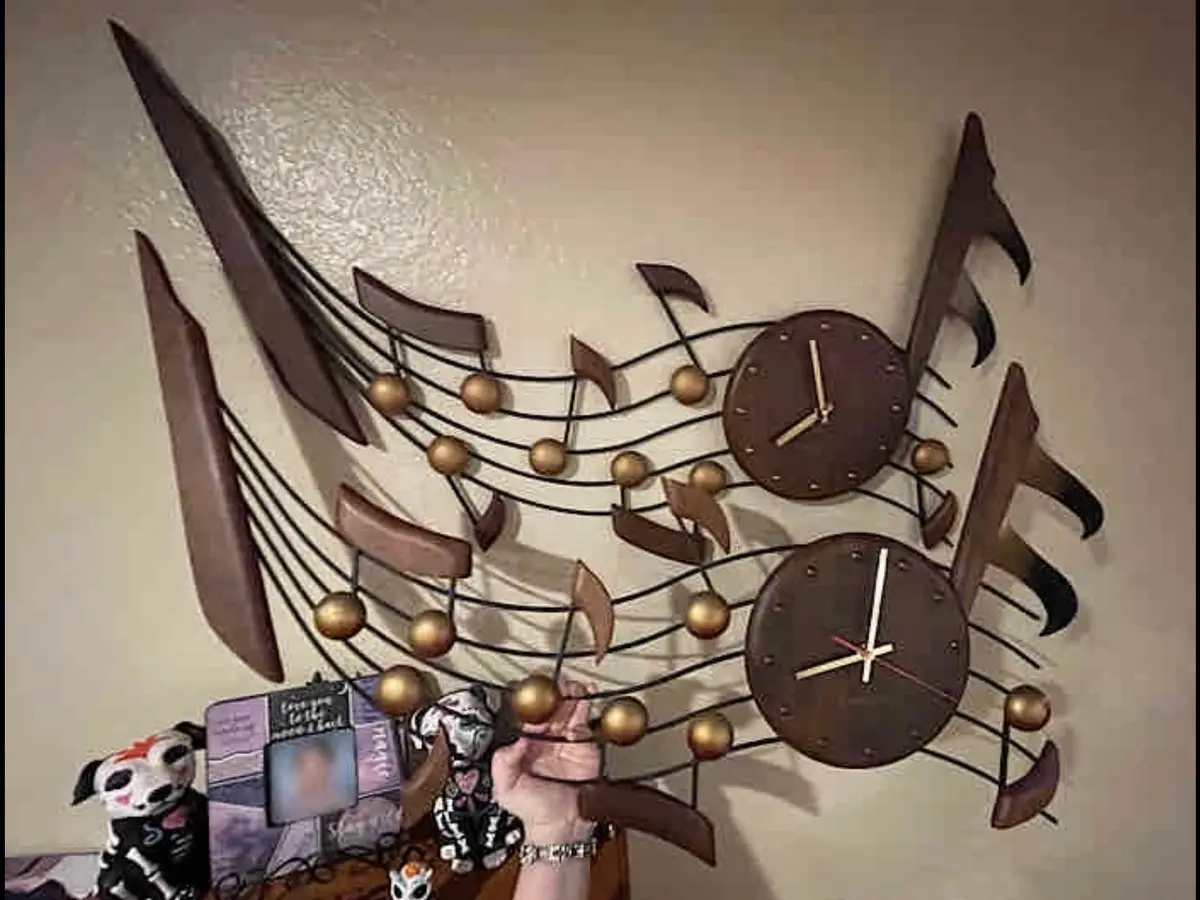 A person's hand reaches up to adjust a large, decorative wall clock shaped like musical notes and staff lines, with two…