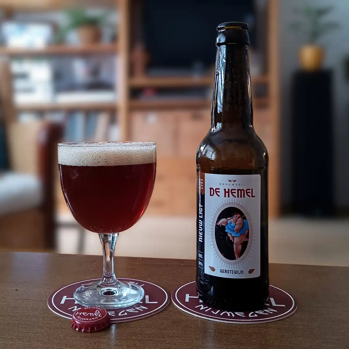 A dark amber beer bottle labeled 'De Hemel' sits next to a filled glass on coasters, with the bottle cap nearby. The label…