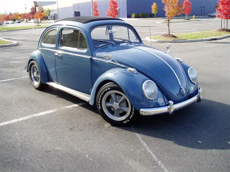 A blue vintage Volkswagen Beetle with chrome trim and whitewall tires is parked in an asphalt lot with white parking lines…